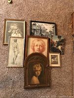 Seven framed and unframed art pieces arranged on carpet displaying variety of portraits, sketches, icon painting, architectural and historical prints
