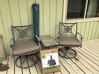 Two metal swivel chairs flanking a tiled top side table, with rolled-up green umbrella behind and boxed umbrella base in front
