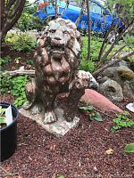 Concrete lion statue sitting on red mulch with visible weathering and moss.