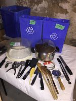 Photo showing two blue recycling bins, Lagostina soup pot, glass cake plate, and various kitchen utensils spread out on a table.