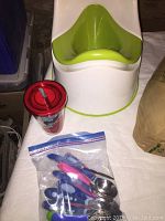 Photo shows green and white potty chair, red plastic cup with straw, and a plastic bag containing colorful plastic clips and metal spoons on white surface.