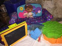 Foam toddler chair featuring Doc McStuffins character and colors with several toys in front including a yellow and green drawing board, blue and red plastic molds, and a green turtle-shaped Play-Doh accessory case.