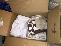 Open cardboard box filled with various bedding items visible including a white comforter and patterned duvet covers and sheets.