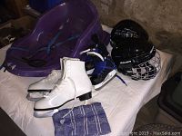 Photo showing purple infant tow sled, two pairs of skates (white women's figure and black toddler hockey), two black helmets (one youth Bauer with cage, one small out-of-date), and blue patterned tea towel.