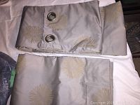 Top view of two folded decorative drapes in light grey with sunburst pattern and silver grommets for hanging.