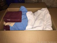 Photo of a cardboard box filled with a mix of towels including white bath towels, a blue towel, burgundy hand towels, and a beige towel.