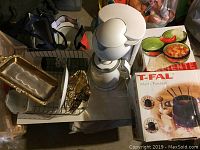 Wide shot of kitchen items arranged on table including T-Fal multi fondue pot box, white Krups coffee maker, dish rack, metal serving trays, water bottle, and bowls.