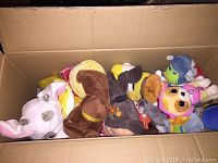 Open cardboard box filled with various small plush toys mostly dogs and other animals, showing multiple colors such as brown, pink, yellow, blue and white.