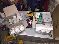 Image showing multiple boxes of assorted lightbulbs including Philips compact fluorescent bulbs and a black box Philips Rough Service outdoor bulb, placed on a white surface with some tools and bags nearby.
