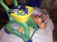 Photo of LeapFrog Pretend & Learn Shopping Cart with a yellow basket and green frame, with a bag of assorted plush toys next to it.