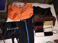 Photo shows seven bags and purses arranged on a white surface. The bright orange Lancaster tote is at the back left. Two Kate Spade small wristlet purses with black, white, and tan color blocking are at the front right. A brown Linda Verdi purse is near the center. A black faux leather Kenzie bag with a tear in the lining, a black yoga mat bag with a blue stripe, and a black Knix Wear fabric bag are also visible.