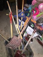 Garden tools piled inside a large plastic bin, showing multiple shovels, hoes, a pitchfork, and other hand tools with wooden and metal handles.