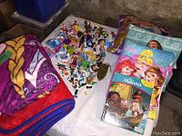 Photo showing three folded Disney themed blankets with Disney princess and Frozen characters, assorted small plastic Disney character toys spread out, two Disney princess pillows, and two soft cloth Disney books including Princess and Moana themes.