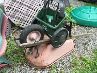 Photo showing a rusty pink metal wheelbarrow with a flat tire and a Scotts green plastic seed spreader with black wheels on gravel ground, surrounded by garden items.