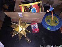 Photo showing gold star tree topper with spiraled base in front of a cardboard box containing toy instruments, small Justice League ornament in red box, and circular cat toy with hanging bird on blue and green base.
