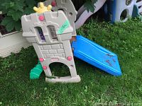 Full view of the Little Tikes castle-themed plastic play structure with blue slide set on grass outdoors. Shows the beige/gray castle frame with pink accents and yellow crown on top plus the blue slide attached on one side.