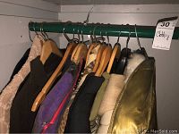 Photo of women's coats and jackets hanging on wooden and metal hangers on a green rail inside a closet showing variety including leather, fur-lined, and fabric coats.