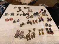 Wide view of over 20 pairs of vintage costume earrings spread on a white cloth, showing various styles and colors including metallics and colorful stones.