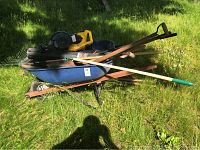 Lot of garden tools outdoors on grass including wheelbarrow, weed trimmer, leaf blower, and hand tools