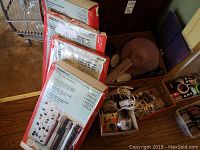 Photo showing four unopened ClosetMaid Deluxe 48-pair shoe organizer boxes with retail labeling, assorted small household items including napkin rings, placemats, kitchen trays, and boxes of cards.