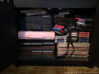 Shelf with stacked LP records, cassette tapes, CDs, and storage containers including zippered cases and plastic boxes.