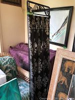 Black decorative screen with detailed white damask patterned black fabric panel, partially visible second purple patterned fabric item behind it, with framed mirror on the floor nearby.