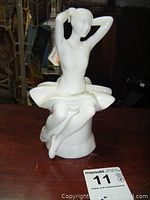 Frontal view of white glossy porcelain ballerina figurine sitting with arms raised behind head, showing crossed legs and tutu over pedestal base.
