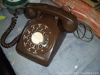 Brown vintage rotary dial telephone front angle showing dial and handset with coiled cord extending to right side, placed on blue surface with background objects.