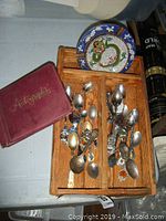 Wooden tray holding vintage collector spoons alongside two autograph books and a cloisonne ashtray