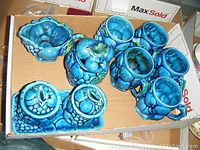 Photo of various blue glazed china items including mugs, cream and sugar set, condiment sets, tea pot, and vase showing vivid blue color and detailed fruit motifs.
