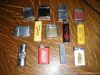 Overview of 13 vintage lighters and 1 lighter cover laid out on wooden surface, showing various styles, brands and colors.