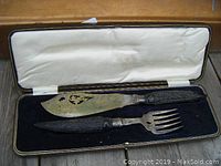 Open case showing carving knife and fork with bone handles and hallmarked sterling bands on a black velvet base and white satin lining.