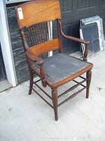 Side angle of antique wooden armchair showing curved top backrest, spindle design, wooden arms, and worn upholstered seat.