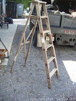 Front angled view of old wooden 6 ft step ladder standing on gravel, showing side steps, wood knots, and aged appearance.