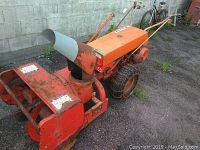 Side view of the orange Gravely two-stage snow blower showing metal chute, auger housing, chain-wrapped rear wheels, paint wear and rust