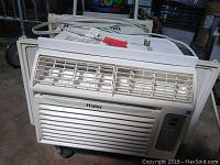 Front angled view of white Haier 7800 BTU window air conditioner with vent louvers, control panel and side window panels on a dolly.