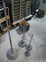Five vintage microphone stands of different sizes standing on concrete floor in a workshop area, metal bases and shafts visible