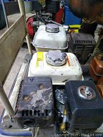 Honda GX120 engine with white fuel tank, black muffler cover, and visible wear and dirt, showing side and top views.