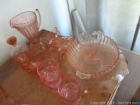 Pink depression glass items on wooden table including pitcher, glasses, bowls, underplates, and decanter with stopper, showing overall lot condition.