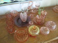 Photo showing the collection of various pink depression glass cups, bowls, jars, drinking glasses, and pitcher. The amberina pressed glass jar is partially visible on the right side.