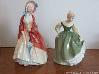Side-by-side image of two Royal Doulton porcelain figurines, one wearing red shawl and bonnet, the other in green dress