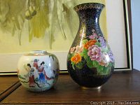 Photo showing two vases: a round Chinese porcelain jar with figures and a taller cloisonné floral vase on a wooden surface.