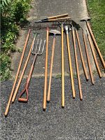 Photo of all garden and lawn tools laid out on driveway showing metal and wooden handled tools including rakes, pitchforks, hoes, pruners, and edgers