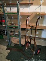 Two heavy duty moving tools: a green appliance dolly and yellow hand truck standing upright indoors on green carpet with shelves in background.