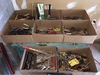 Six cardboard boxes and a plastic crate on a workbench, filled with various hand tools including screwdrivers, wrenches, hammers, pliers, bits, punches, wire cutters, tin snips, and small tools.