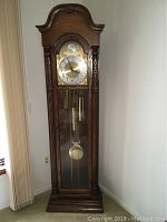 Full front view of oak case grandfather clock showing bonnet pediment, glass door, dial, weights and pendulum