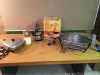 Photo showing all small kitchen appliances on a table including the Sunbeam electric fry pan, OSKAR food chopper, Hamilton Beach stand mixer, LeChef food processor box, and Farberware indoor grill.
