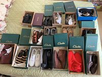 Overview photo showing 17 pairs of women's shoes in their boxes on a bed, mostly Clarks brand, including boots, sandals, and shoes in various colors like black, brown, tan, white, and red.