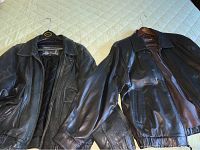 Two black leather men's jackets laid out on bed, showing full front view with collars and sleeves extended.