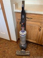 Full view of upright vacuum against wood cabinetry showing handle, cyclone bin and ball base